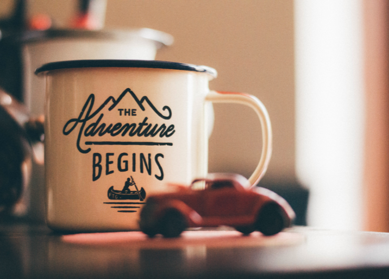 mug with a mountain graphic and the words "Adventure Begins" on the side. toy car in front of the mug on a table