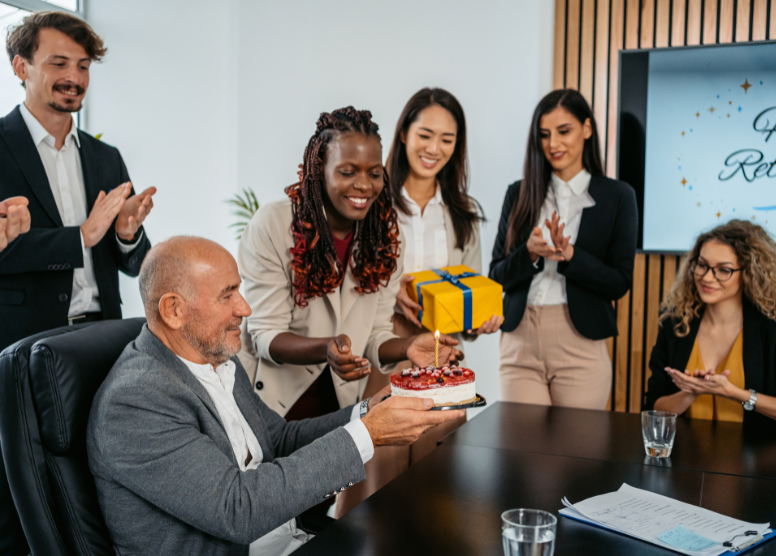man sitting at a table holding a cake. coworkers standing around him smiling and clapping