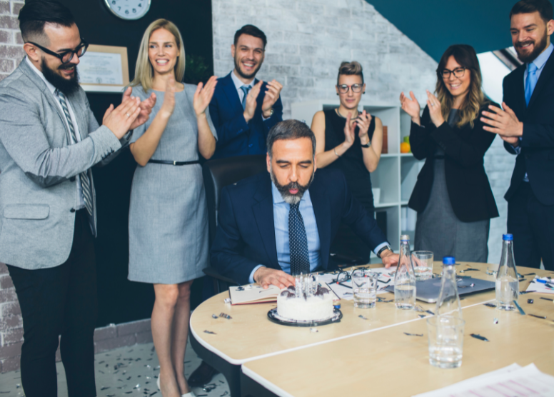 people in an office celebrating a coworker's retirement. Man sitting at a table blowing out candles on a cake; people standing behind him clapping